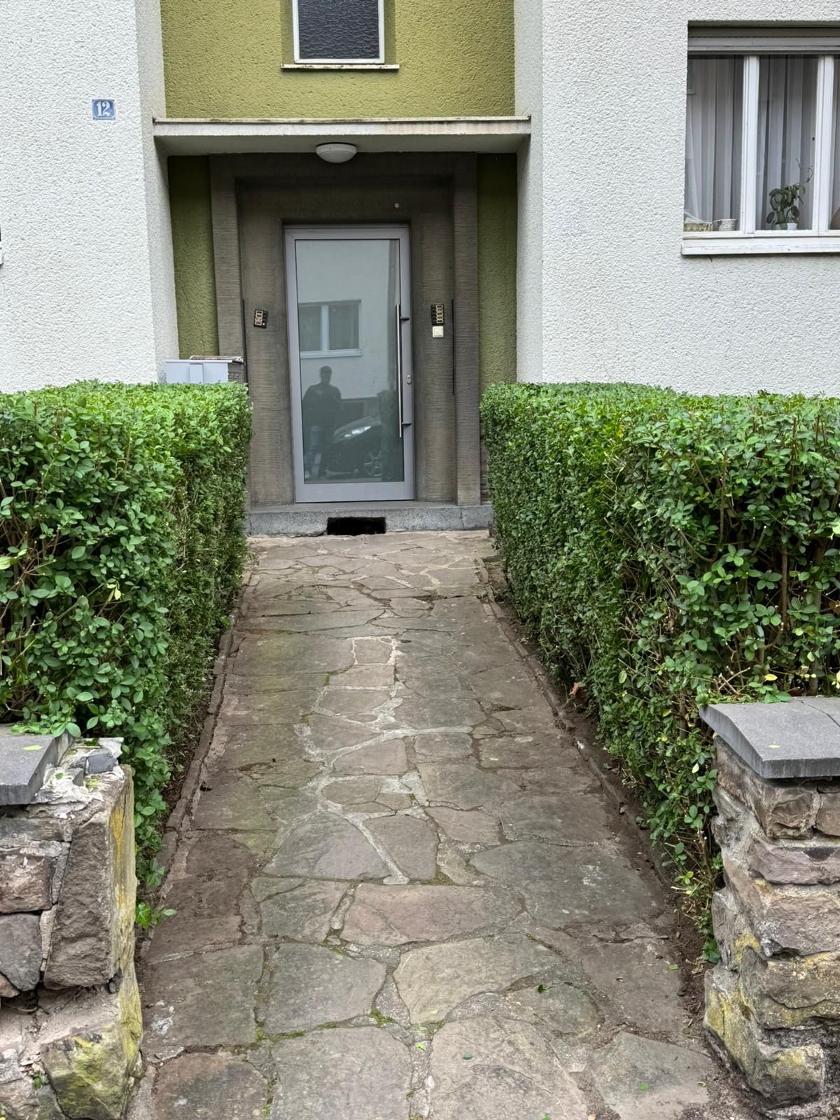 Ein Mann steht mit neutralem Gesichtsausdruck hinter einer Glastür vor grüner Fassade
