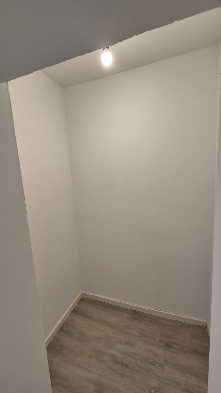 Ein leerer, kleiner Raum mit weißen Wänden, grauem Holzboden und einer Glühbirne an der Decke