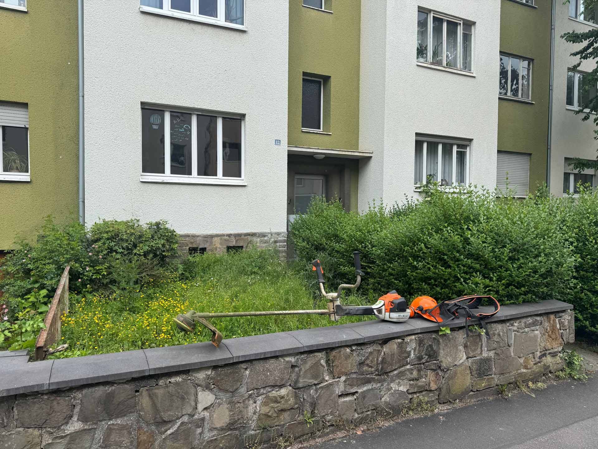 Vor einem grünen Wohnhaus liegt eine Motorsense mit Helm und Schutzbrille auf einer Mauer