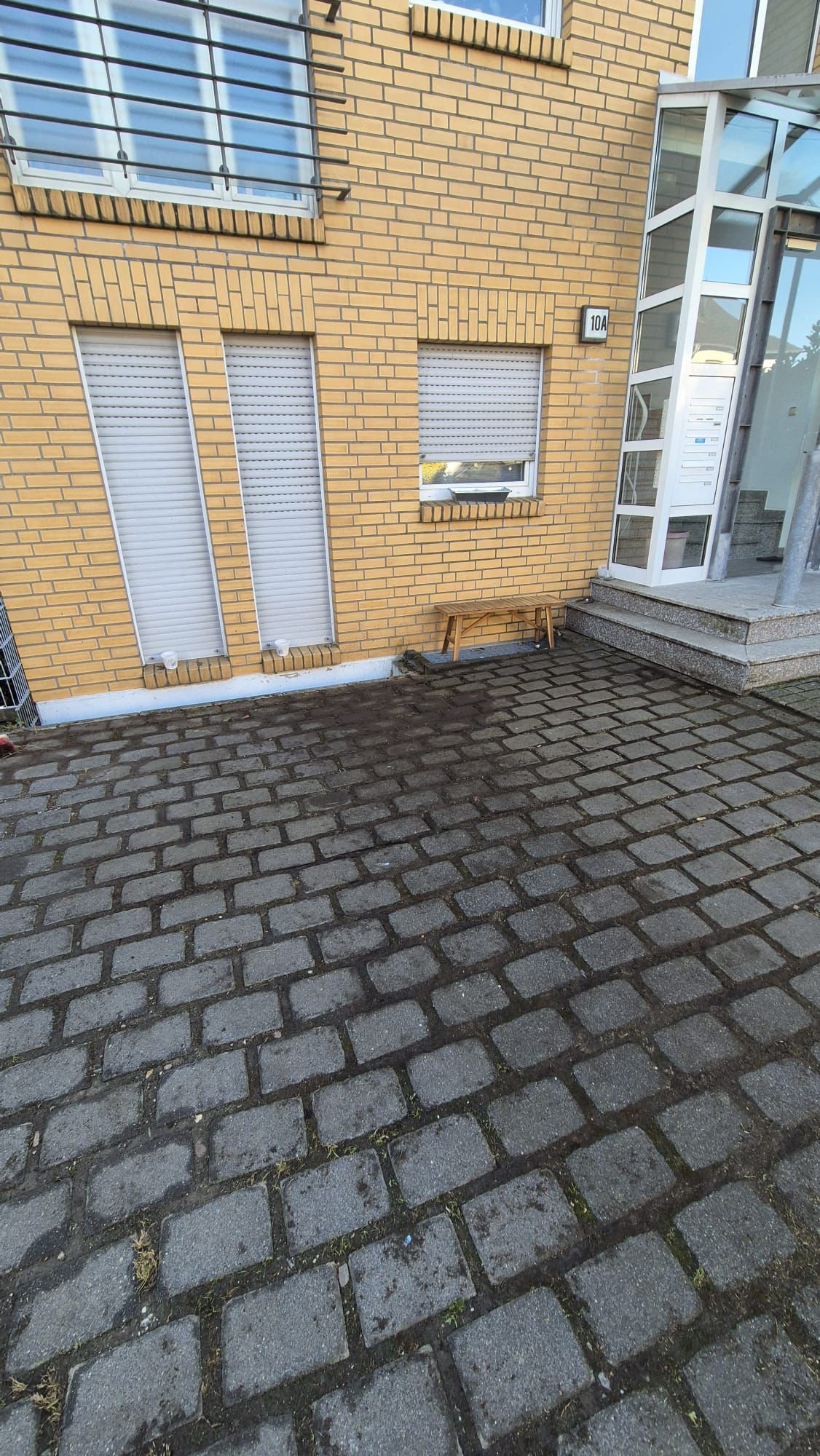 Gelbes Backsteingebäude mit geschlossenen Rollläden, gläsernem Eingang und nassen Pflastersteinen